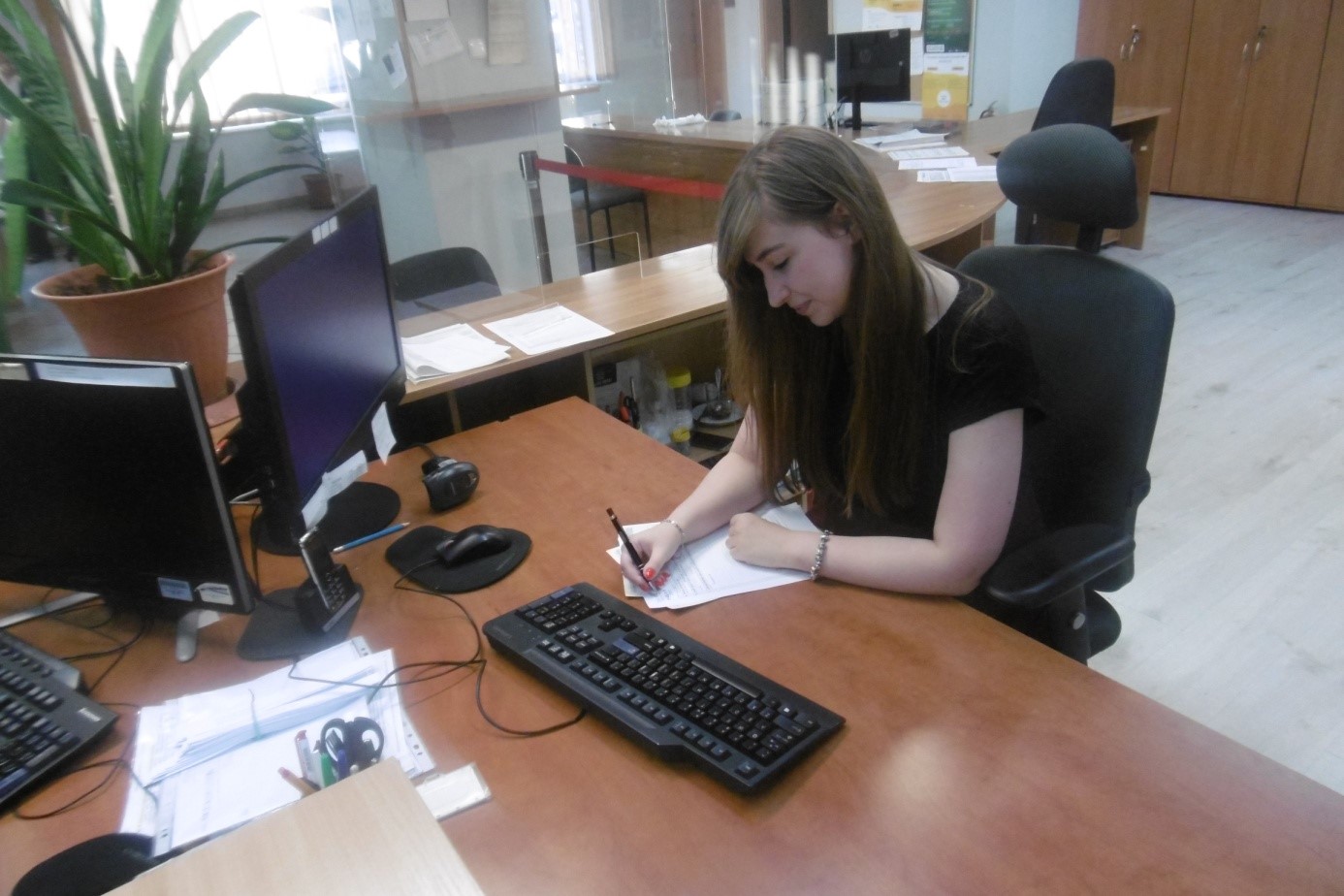 Wnętrze urzędu, przy biurku siedzi młoda kobieta i wypełnia dokumenty