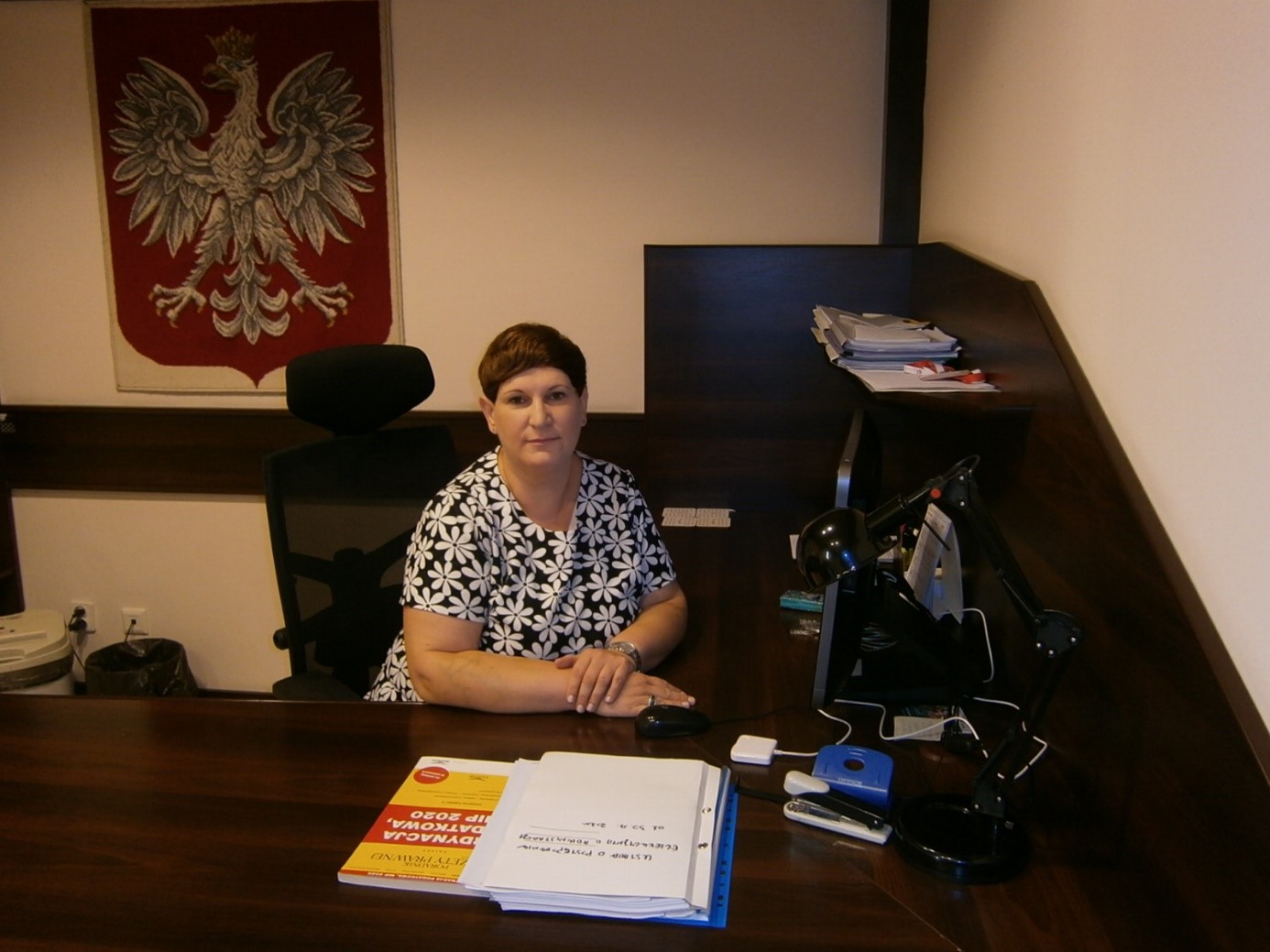 Wnętrze urzędu, przy biurku z dokumentami siedzi kobieta, patrzy w obiektyw, za nią wisi duże godło Polski 
