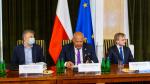 Konferencja prasowa Ministerstwa Finansów w sprawie budżetu na 2021. Przy stole n atle flag Unii Europejskiej i Polski przy stole siedzi trzech panów.