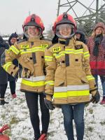 Dwie uśmiechnięte młode kobiety stoją w strojach strażackich. W tle zaśnieżony trawnik.