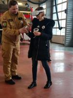 Kobieta ubrana w specjalny strój VR - na głowie specjalne gogle, a w rękach dżojstik. Obok strażak pomagający jej w obsłudze urządzenia.