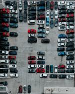 na betonowym parkingu stoi kilkadziesiąt samochodów osobowych, większość w kolorze: białe, czerwone, szare i czarne.