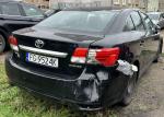 Tył czarnej Toyoty Avensis sedan z uszkodzonym błotnikiem oraz zderzakiem oklejonym srebrną taśmą, widoczne wgniecenie na zderzaku oraz liczne zarysowania lakieru
