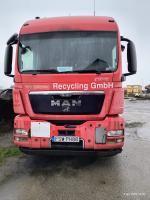 Przód samochodu ciężarowego  MAN TTGS 26.440. Kabina koloru czerwonego. Lusterka boczne koloru czarnego. Grill centralny w kolorze  czarnym ze srebrnym logo marki i napisem MAN. Zderzak czerwony. Dolna blenda zderzaka koloru czarnego. Na masce zarysowania i uszkodzenia lakieru. Na mace, po prawej stronie pomiędzy szybą a grillem, napis koloru białego o treści „Recycling GmbH