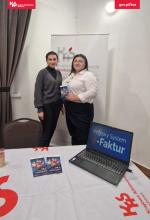 sala, w rogu stoi baner reklamowy Izby Administracji Skarbowej w Zielonej Górze, na stole leży obrus z logo KAS, na stole leżą ulotki, laptop i balony, przed banerem stoją dwie pracownice US