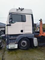 lewy  przód samochodu ciężarowego marki MAN TGA   koloru białego z widocznymi odpryskami lakieru na drzwiach i tyle kabiny . Lusterka i klamka koloru czarnego. Próg nadkola czarny.  Koła na srebrnych, stalowych felgach. Rama  i mechanizm hakowy w kolorze  pomarańczowym