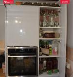 Biała szafka z piekarnikiem. W otwartej szafce widoczne liczne butelki szklane i plastikowe.